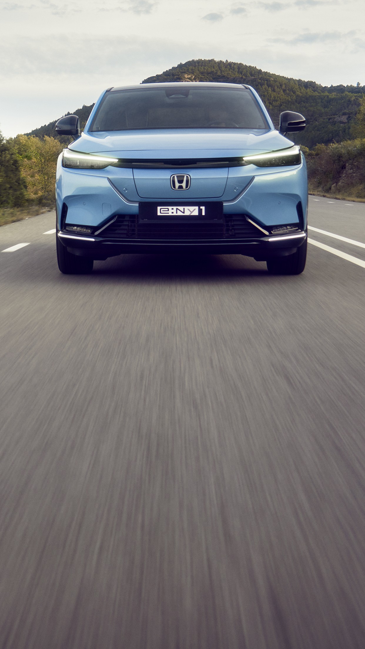 Honda e:Ny1 i Aqua Topaz Metallic kører på en snoet vej med grønne bakker i baggrunden.