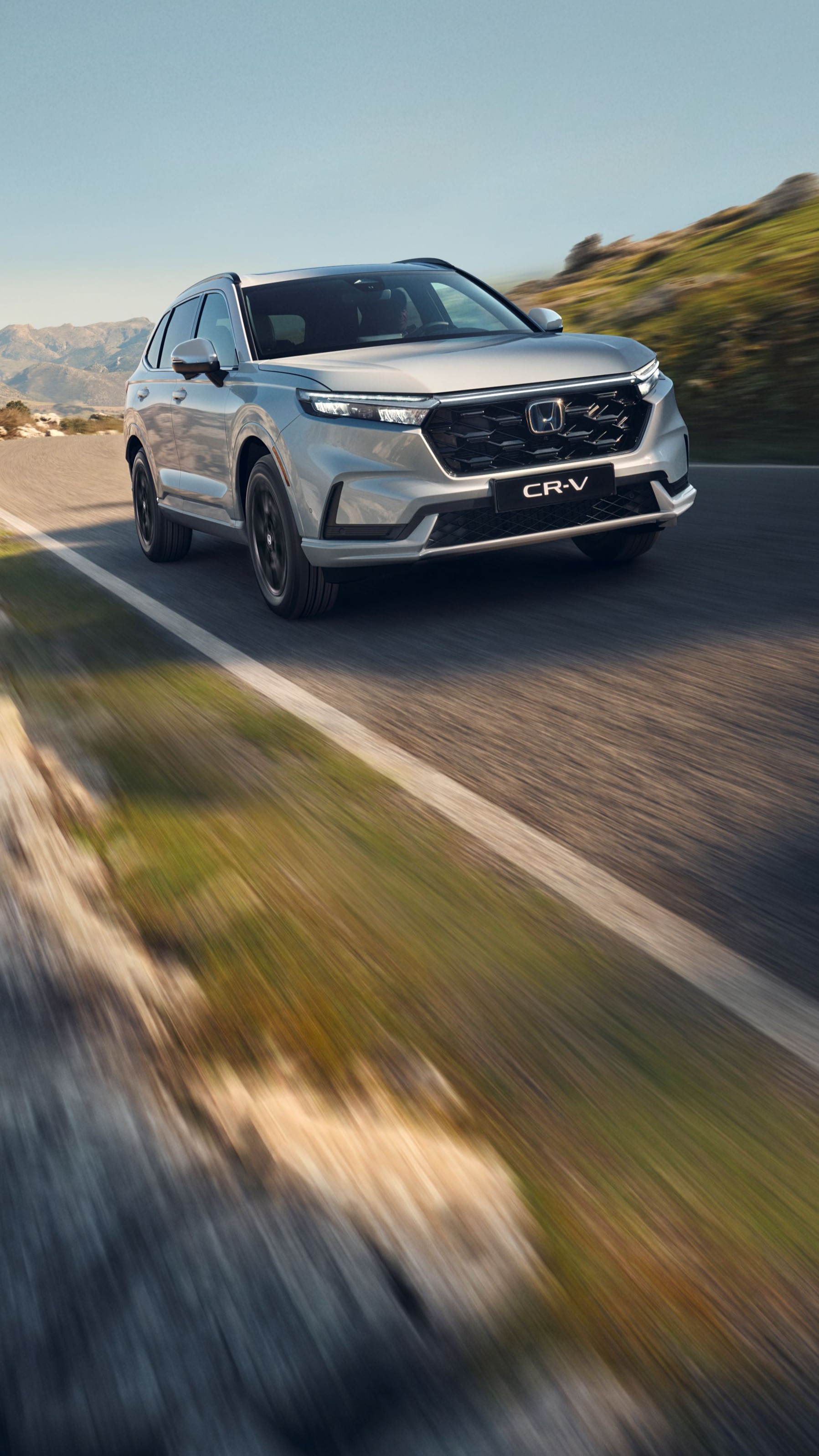 En sølvfarvet Honda CR-V SUV kører på en snoet bjergvej med klippefyldt terræn og bjerge i baggrunden. Køretøjet er i bevægelse, hvilket skaber en følelse af fart og eventyr i det rå landskab.