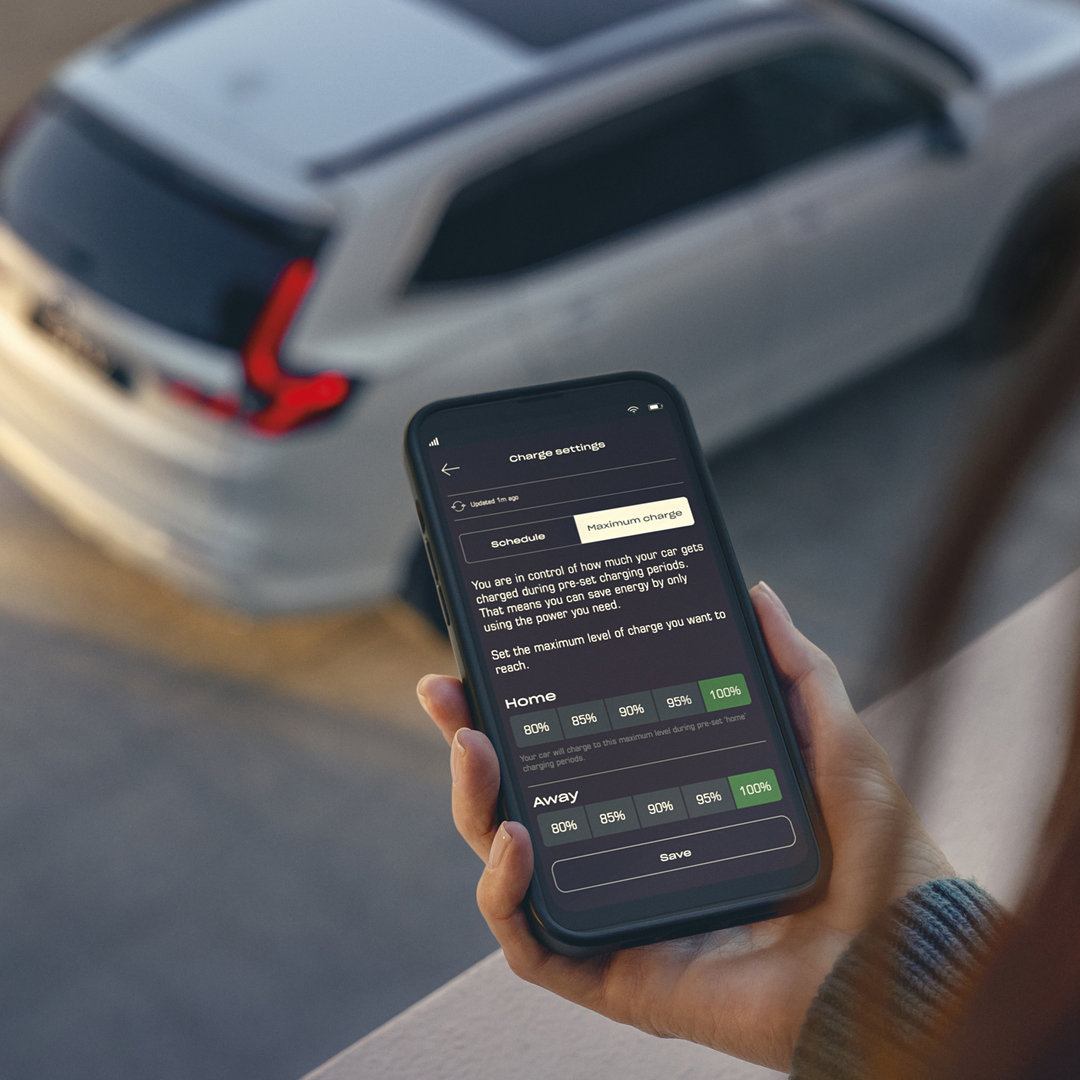 En person holder en smartphone, der viser en app til opladningsindstillinger for køretøjet, mens en hvid SUV holder parkeret i baggrunden.