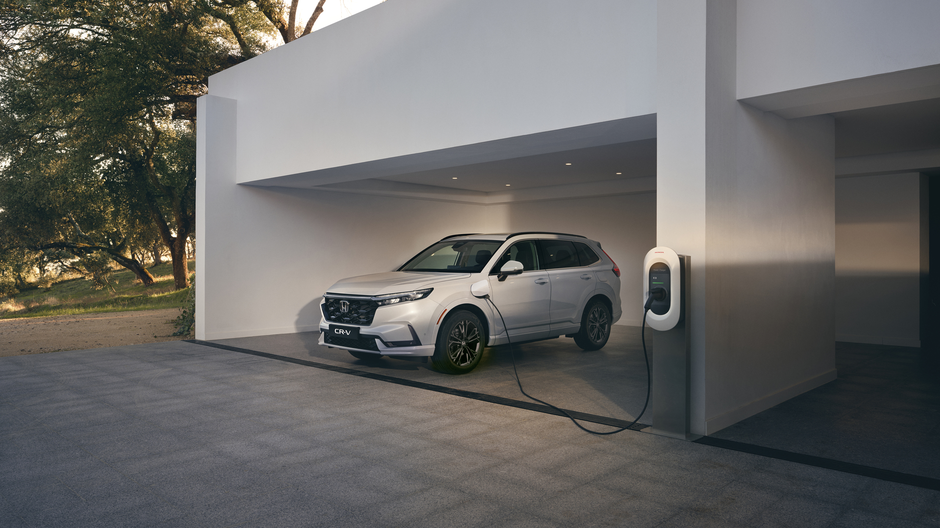 En hvid Honda CR-V plug-in hybrid står i en minimalistisk hvid carport, tilsluttet en vægmonteret oplader til elektrisk køretøj.