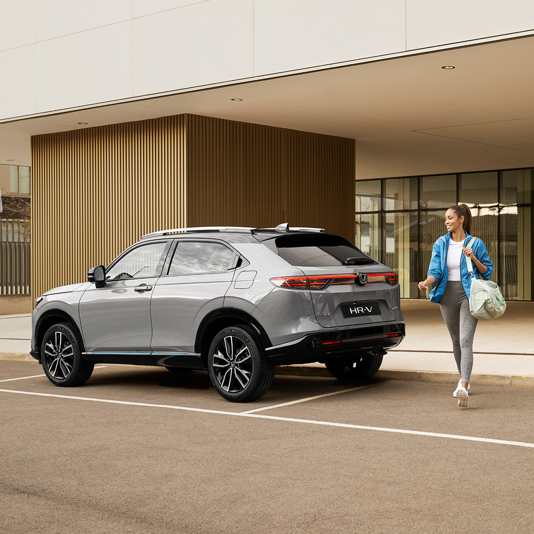 En sølvfarvet Honda HR-V parkeret foran en moderne bygning, mens en kvinde går forbi med en taske, hvilket signalerer urban hverdagsbrug og bekvemmelighed.