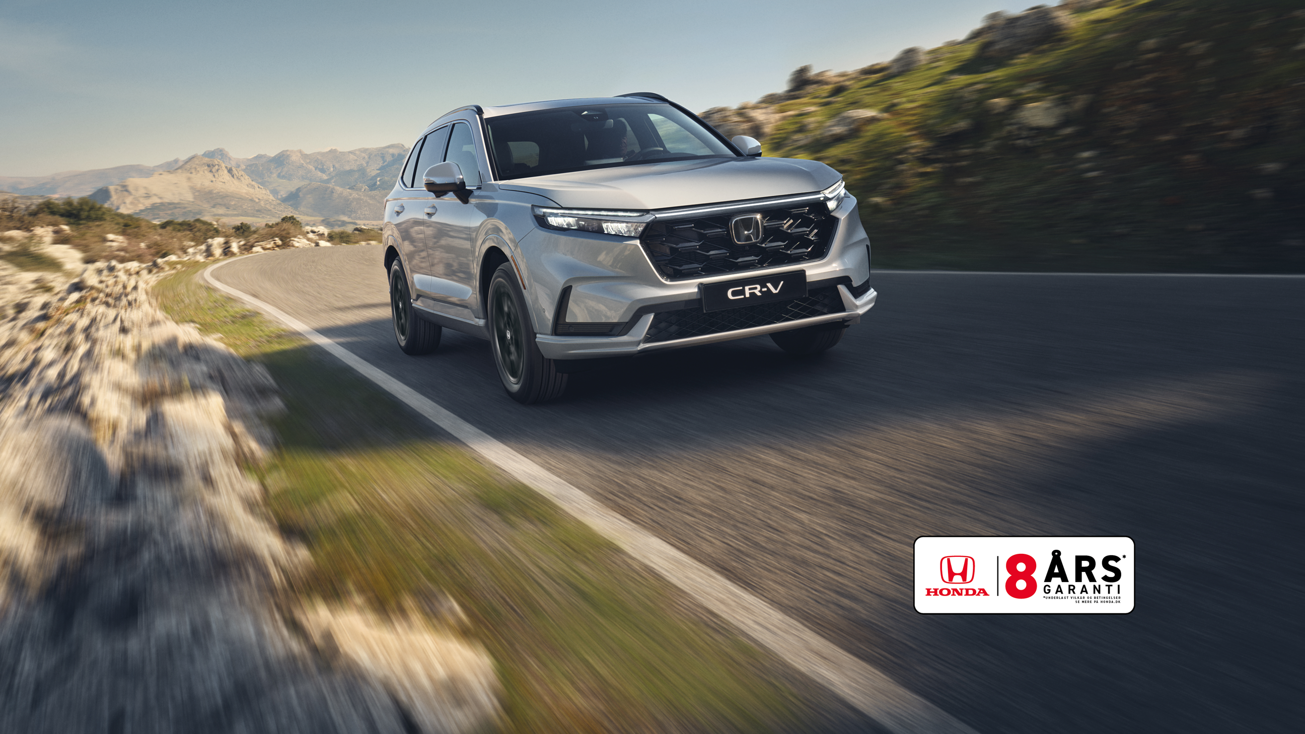 En sølvfarvet Honda CR-V SUV kører på en snoet bjergvej med klippefyldt terræn og bjerge i baggrunden. Køretøjet er i bevægelse, hvilket skaber en følelse af fart og eventyr i det rå landskab.