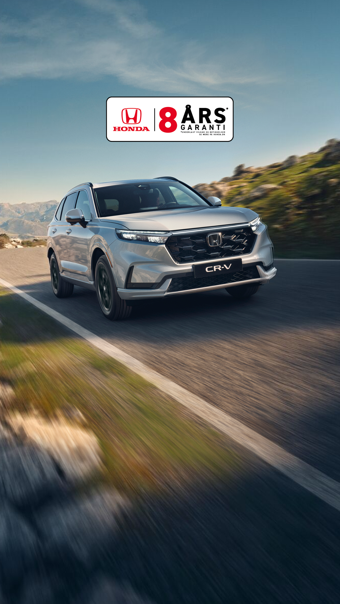 En sølvfarvet Honda CR-V SUV kører på en snoet bjergvej med klippefyldt terræn og bjerge i baggrunden. Køretøjet er i bevægelse, hvilket skaber en følelse af fart og eventyr i det rå landskab.
