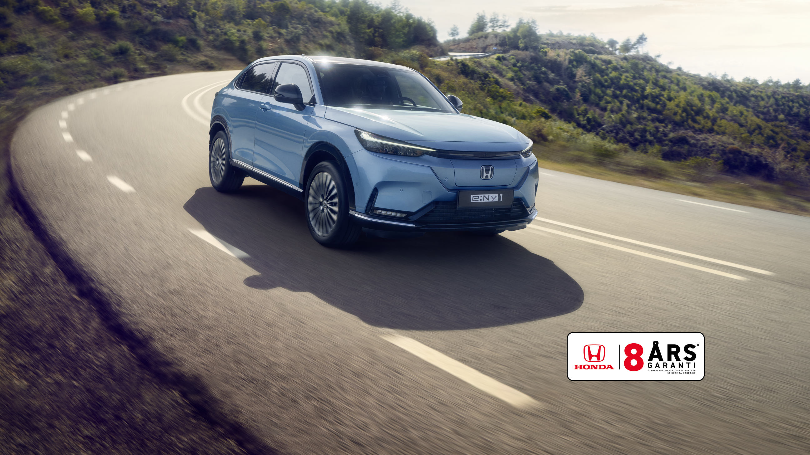 Honda e:Ny1 i Aqua Topaz Metallic kører på en snoet vej med grønne bakker i baggrunden.
